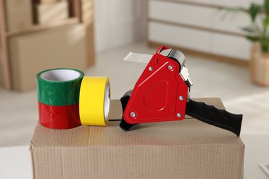 Photo of Dispenser with adhesive tape rolls on cardboard box indoors, closeup