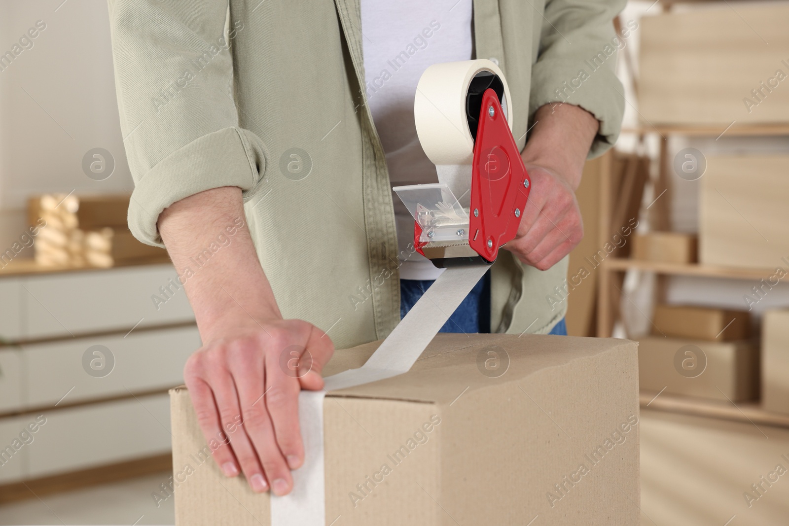 Photo of Man using dispenser with adhesive tape to seal cardboard box indoors, closeup
