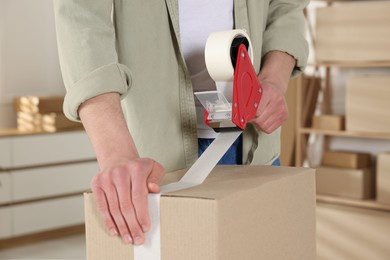 Photo of Man using dispenser with adhesive tape to seal cardboard box indoors, closeup