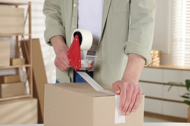 Photo of Man using dispenser with adhesive tape to seal cardboard box indoors, closeup