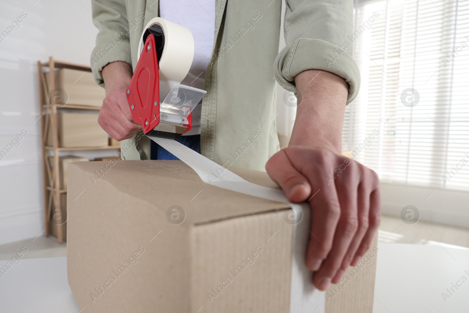 Photo of Man using dispenser with adhesive tape to seal cardboard box indoors, closeup