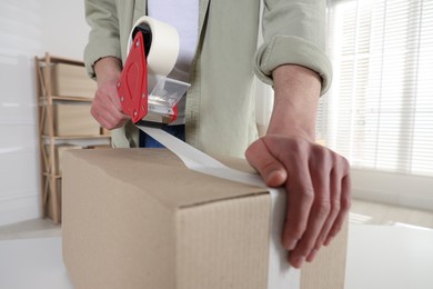 Photo of Man using dispenser with adhesive tape to seal cardboard box indoors, closeup