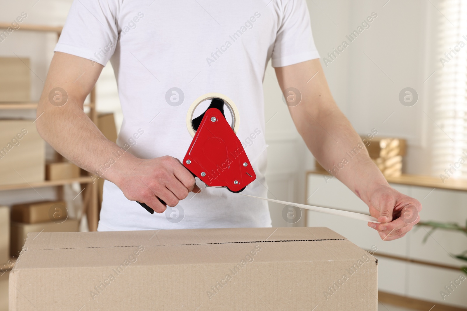 Photo of Man using dispenser with adhesive tape to seal cardboard box indoors, closeup