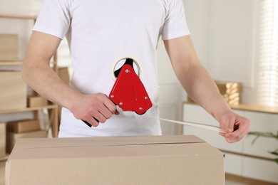 Photo of Man using dispenser with adhesive tape to seal cardboard box indoors, closeup