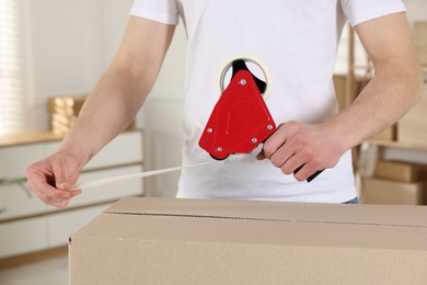 Photo of Man using dispenser with adhesive tape to seal cardboard box indoors, closeup