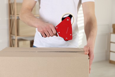 Photo of Man using dispenser with adhesive tape to seal cardboard box indoors, closeup