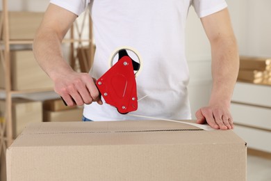 Photo of Man using dispenser with adhesive tape to seal cardboard box indoors, closeup