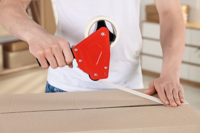Photo of Man using dispenser with adhesive tape to seal cardboard box indoors, closeup