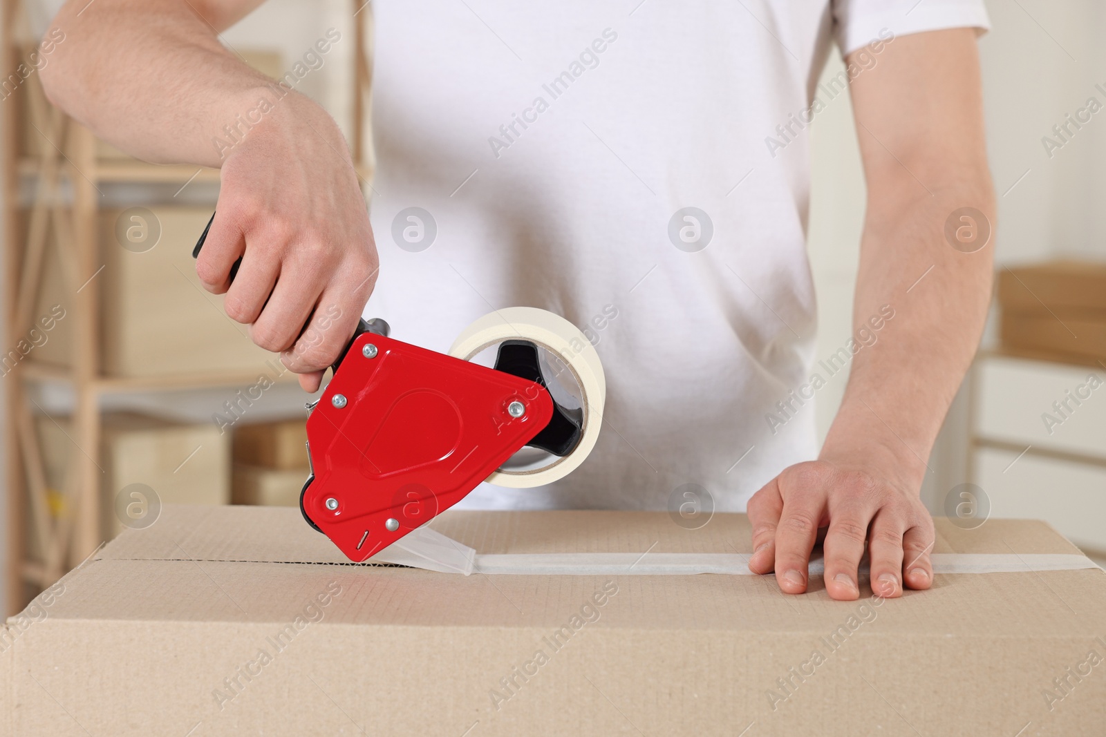 Photo of Man using dispenser with adhesive tape to seal cardboard box indoors, closeup
