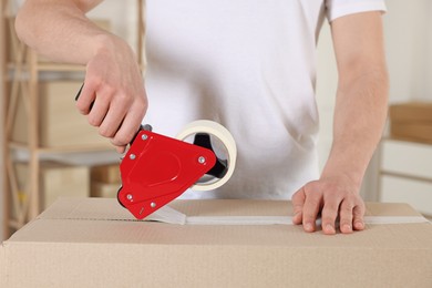 Photo of Man using dispenser with adhesive tape to seal cardboard box indoors, closeup
