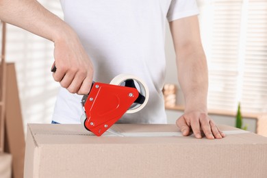 Photo of Man using dispenser with adhesive tape to seal cardboard box indoors, closeup