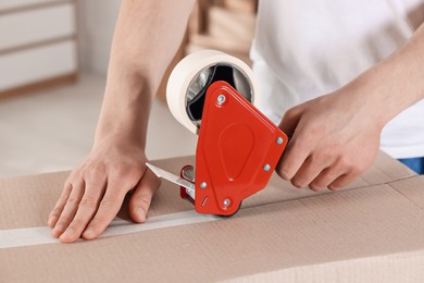 Photo of Man using dispenser with adhesive tape to seal cardboard box indoors, closeup