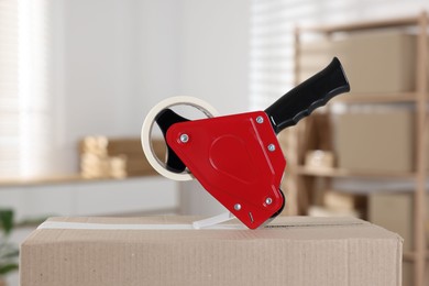 Photo of Dispenser with adhesive tape on cardboard box indoors, closeup