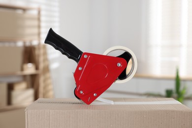 Photo of Dispenser with adhesive tape on cardboard box indoors, closeup
