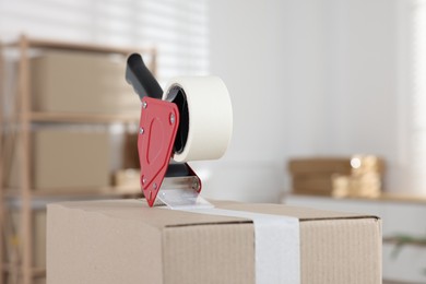 Photo of Dispenser with adhesive tape on cardboard box indoors, closeup