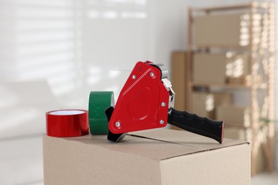 Photo of Dispenser with adhesive tape rolls on cardboard box indoors, closeup