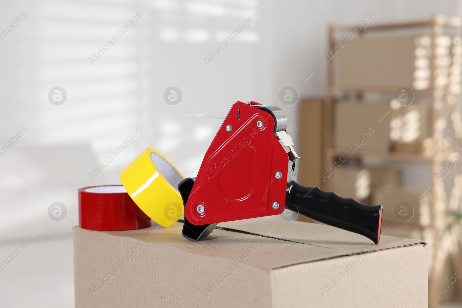 Photo of Dispenser with adhesive tape rolls on cardboard box indoors, closeup