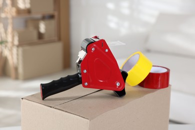 Photo of Dispenser with adhesive tape rolls on cardboard box indoors, closeup