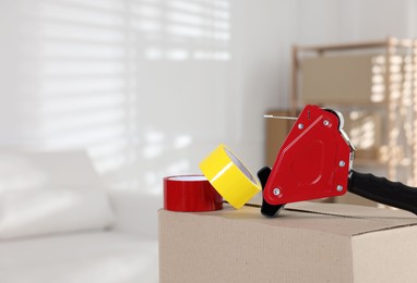 Photo of Dispenser with adhesive tape rolls on cardboard box indoors, closeup