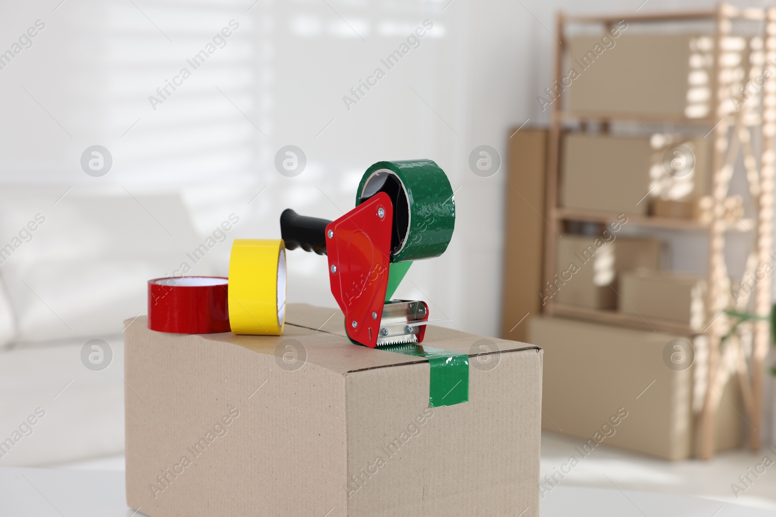 Photo of Dispenser with adhesive tape rolls on cardboard box indoors, closeup