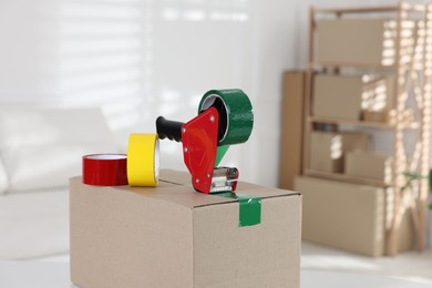 Photo of Dispenser with adhesive tape rolls on cardboard box indoors, closeup