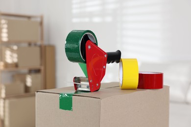Photo of Dispenser with adhesive tape rolls on cardboard box indoors, closeup