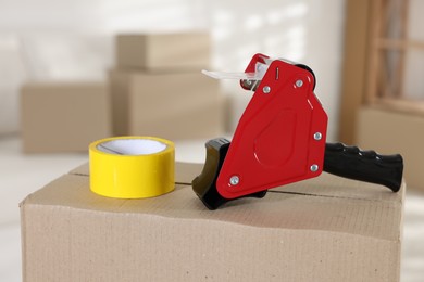 Photo of Dispenser with adhesive tape on cardboard box indoors, closeup