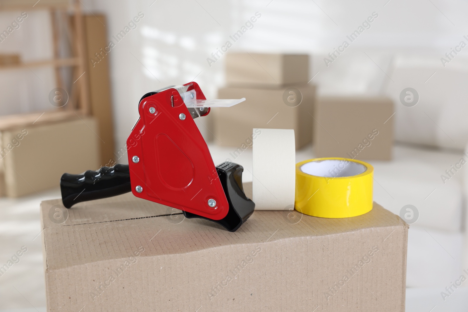 Photo of Dispenser with adhesive tape rolls on cardboard box indoors, closeup