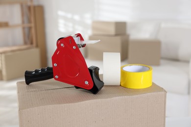 Photo of Dispenser with adhesive tape rolls on cardboard box indoors, closeup