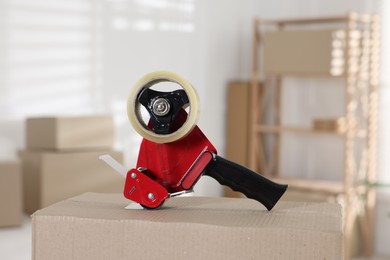 Photo of Dispenser with adhesive tape on cardboard box indoors, closeup