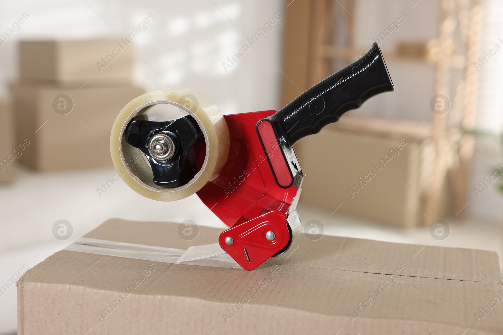 Photo of Dispenser with adhesive tape on cardboard box indoors, closeup