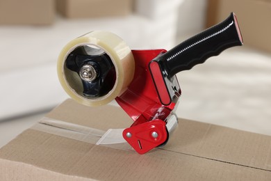 Photo of Dispenser with adhesive tape on cardboard box indoors, closeup
