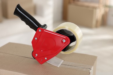 Photo of Dispenser with adhesive tape on cardboard box indoors, closeup