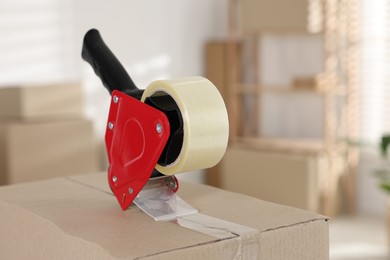 Photo of Dispenser with adhesive tape on cardboard box indoors, closeup