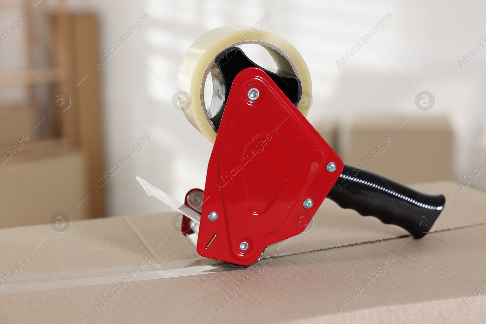 Photo of Dispenser with adhesive tape on cardboard box indoors, closeup