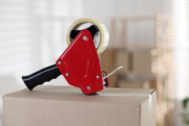 Photo of Dispenser with adhesive tape on cardboard box indoors, closeup