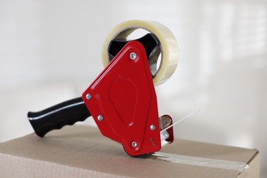 Photo of Dispenser with adhesive tape on cardboard box indoors, closeup