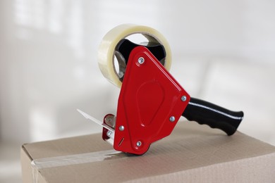 Photo of Dispenser with adhesive tape on cardboard box indoors, closeup