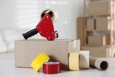 Photo of Dispenser with adhesive tape rolls and cardboard box on desk indoors