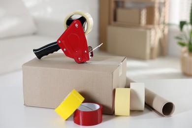 Photo of Dispenser with adhesive tape rolls and cardboard box on desk indoors
