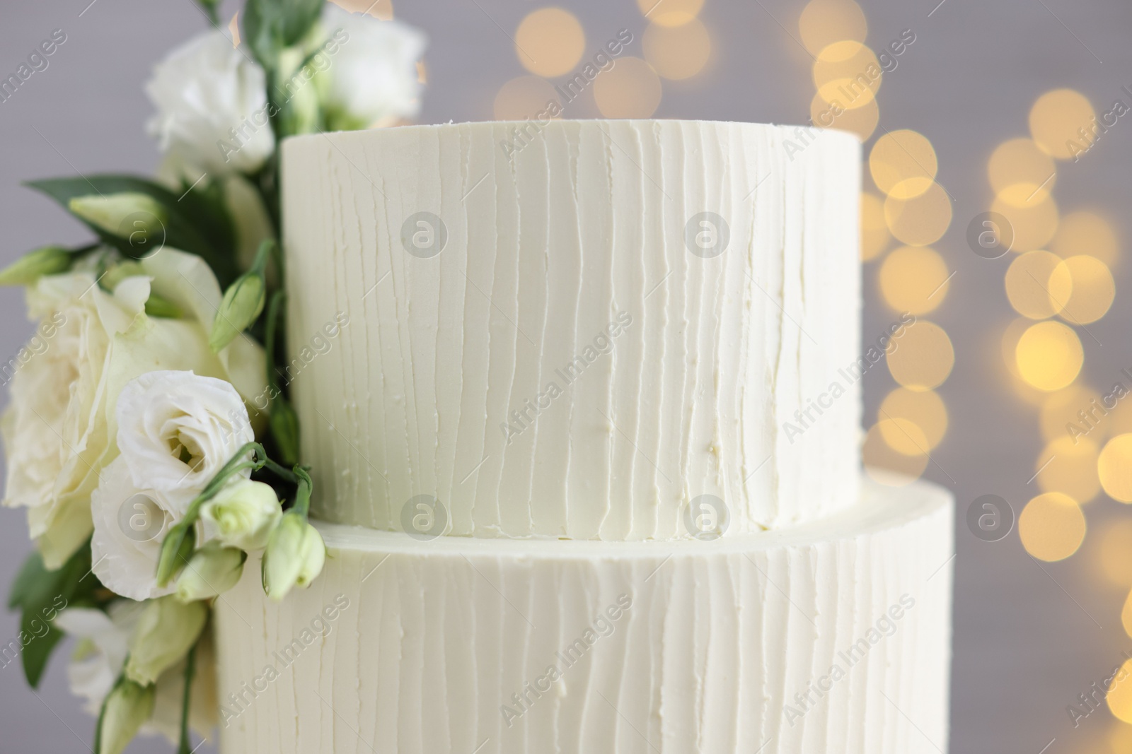 Photo of Delicious wedding cake decorated with flowers on grey background with blurred lights, closeup