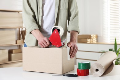 Photo of Man using dispenser with adhesive tape to seal cardboard box at desk indoors, closeup