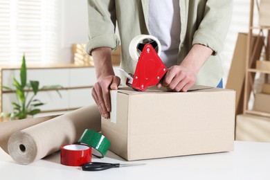 Photo of Man using dispenser with adhesive tape to seal cardboard box at desk indoors, closeup