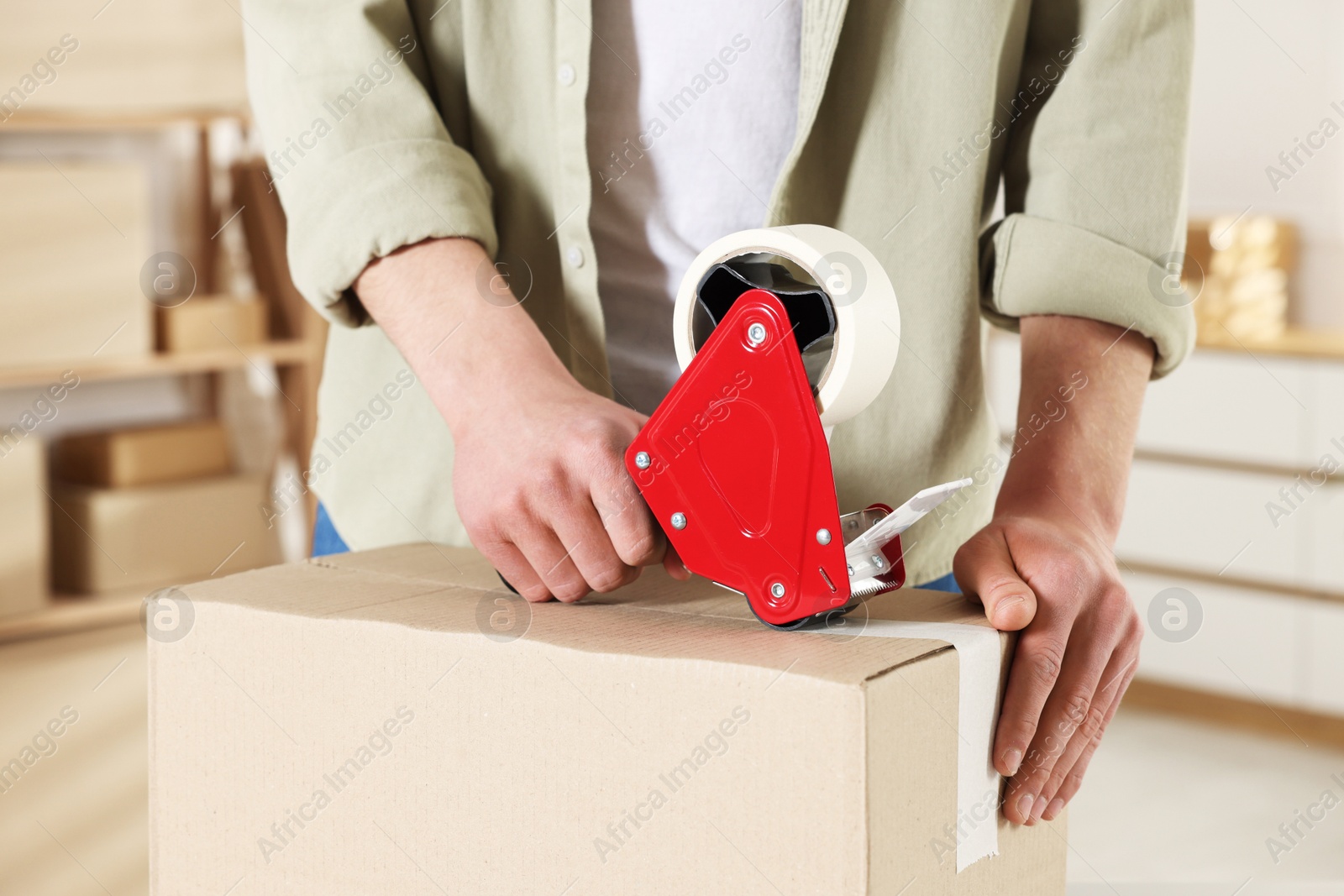 Photo of Man using dispenser with adhesive tape to seal cardboard box indoors, closeup