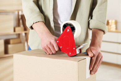 Photo of Man using dispenser with adhesive tape to seal cardboard box indoors, closeup