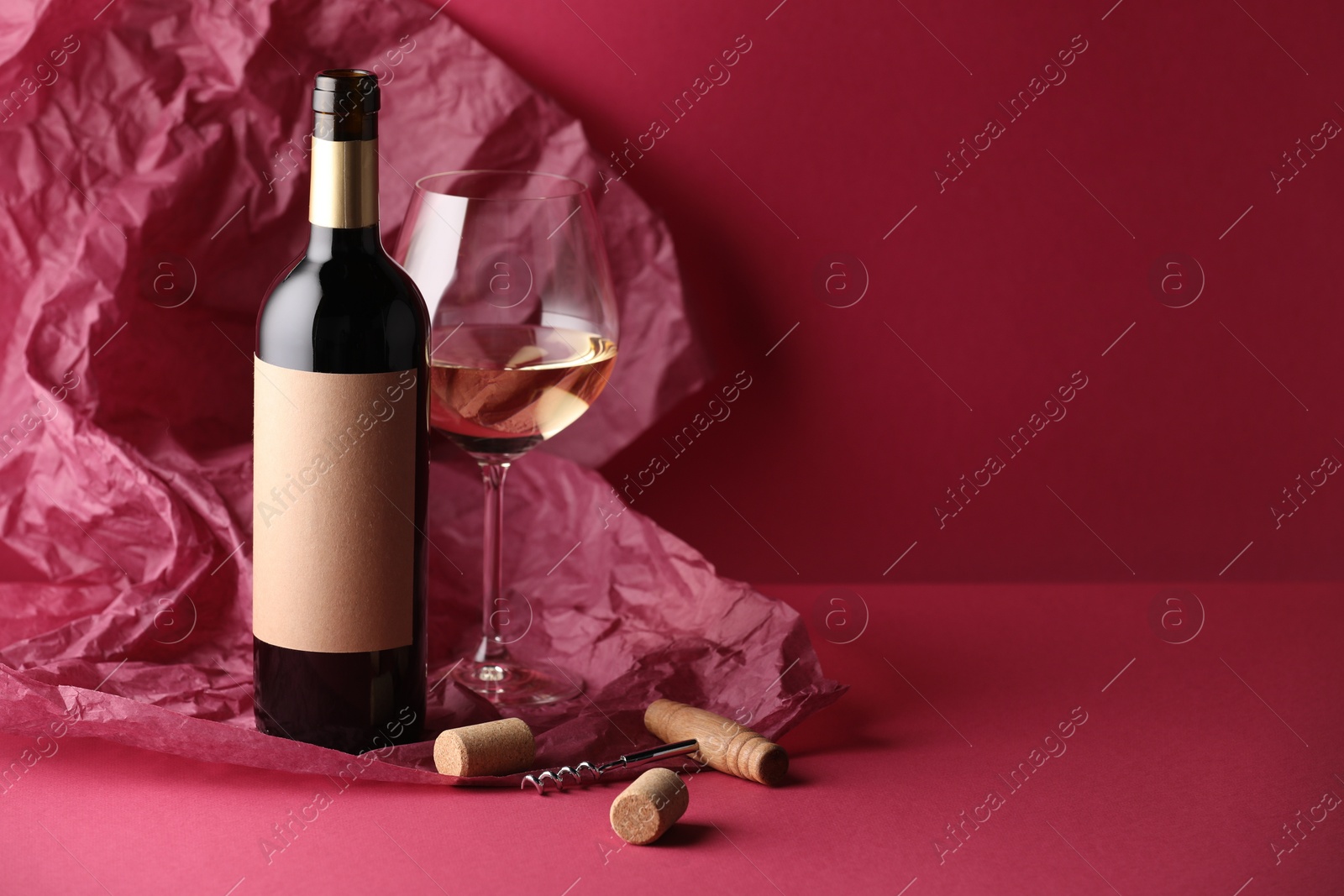 Photo of Bottle and glass of wine with corkscrew, corks and paper on crimson background. Space for text