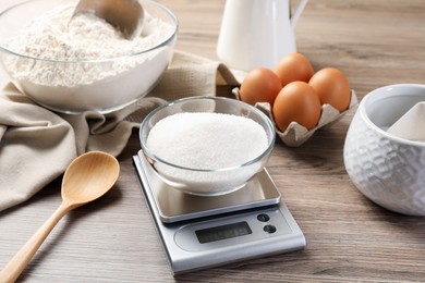 Photo of Digital kitchen scale with bowl of sugar among other products on wooden table