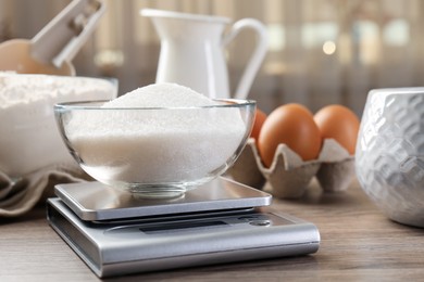 Photo of Digital kitchen scale with bowl of sugar among other products on wooden table, closeup