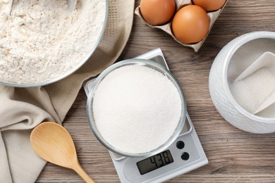 Photo of Digital kitchen scale with bowl of sugar among other products on wooden table, flat lay