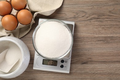 Photo of Digital kitchen scale with bowl of sugar and eggs on wooden table, flat lay